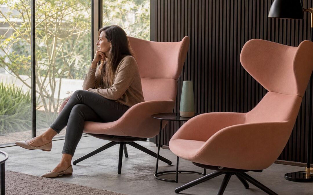 Woman sitting in a dusty rose wingback swivel chair looking out a window in a modern living room with dark wood paneling.